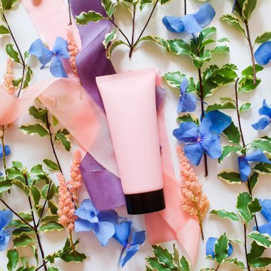 Siyah kapaklı pembe plastik tüp, taze yeşiller, mavi ve pembe çiçekler, beyaz arka planda ipek kurdeleler. Damgalamak ve etiketlemek için bir şişe. Doğal organik kaplıca konsepti.