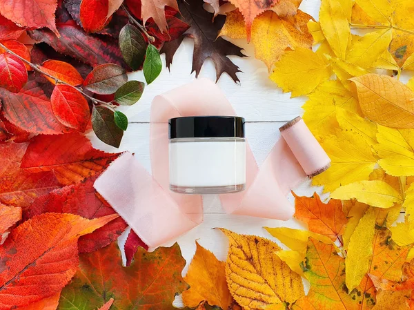 Mockup of facial or eye cream in white glass jar with black cap, pink silk ribbon and autumn bright colorful leaves on white wooden table. Top view. Natural organic cosmetics concept. Fall flatlay