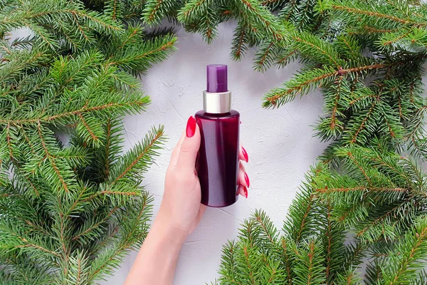 Kahverengi sprey şişenin markasız maketi, kırmızı tırnaklı beyaz kadın elleri ve gri masanın üzerinde Noel ağacı dalları. Doğal organik spa kozmetik konsepti. Düz yaşam biçimi.