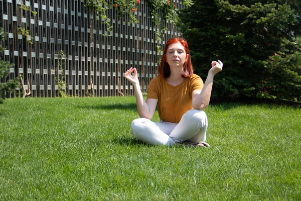 Uma mulher com um rosto feliz. A nacionalidade europeia. Em roupas do dia a dia. Com cabelo ruivo, sentado na posição de Lótus. No parque na grama em um dia ensolarado. Verão . — Fotografia de Stock