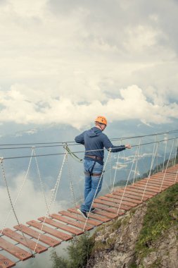 Güvenlik ağı takan bir erkek ve bir kadın. Uçurumun üzerinde ip bir köprüde yürüyorlar. Dağlardaki asma köprüden geçmek. Olağanüstü bir spor. Arkadan manzara. Bulutların arka planında