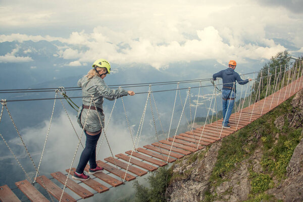 A man and a woman wearing a safety net. They are walking on a rope bridge over an abyss. Crossing a suspension bridge in the mountains. Extreme sport. The view from the back. On a background of clouds