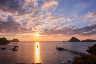 Güneşle aynı hizada olan bir yelkenli teknesi Labuan Bajo 'da bir marinayla birlikte hafif bulutlu bir gün batımında suya vuruyor.
