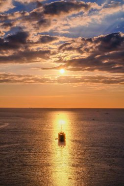Güneşe doğru giden bir yelkenli. Güneşle aynı hizada. Hafif bulutlu bir günbatımında suya yansıyor.