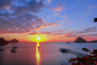 Güneşle aynı hizada olan bir yelkenli teknesi Labuan Bajo 'da bir marinayla birlikte hafif bulutlu bir gün batımında suya vuruyor.