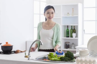 Genç Asyalı kadın mutfakta durup mutfağı temizliyor ve kameraya gülümsüyor.