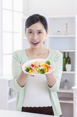 Young Asian woman standing in the kitchen holding a plate of vegetables and smiling at the camera