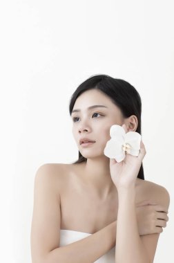 Genç Asyalı kadın başının yanında bir çiçek tutuyor ve başka tarafa bakıyor.,