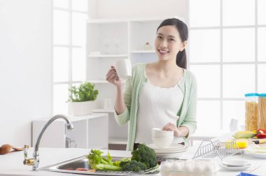 Genç Asyalı kadın mutfakta durup mutfağı temizliyor ve kameraya gülümsüyor.