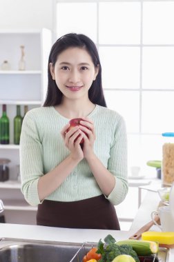 Mutfakta bir elma tutarak ve kameraya gülümseyen duran Genç Asyalı kadın