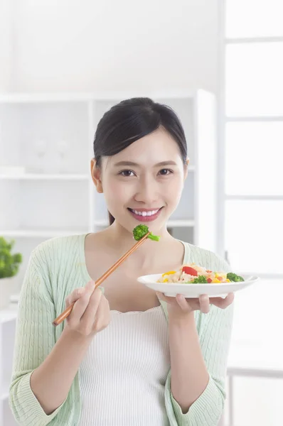 年轻的亚洲女人站在厨房里 端着盘子吃饭 微笑着看着摄像机 — 图库照片