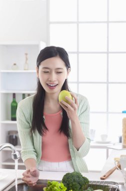 Genç Asyalı kadın mutfakta bir elma yeme ve uzağa arıyorsunuz gülümseyen ayakta