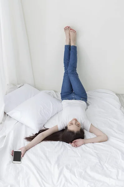 笑みを浮かべて彼女の携帯電話を保持している彼女の足を入れてベッドに横になっているジーンズの若いアジア女性 — ストック写真