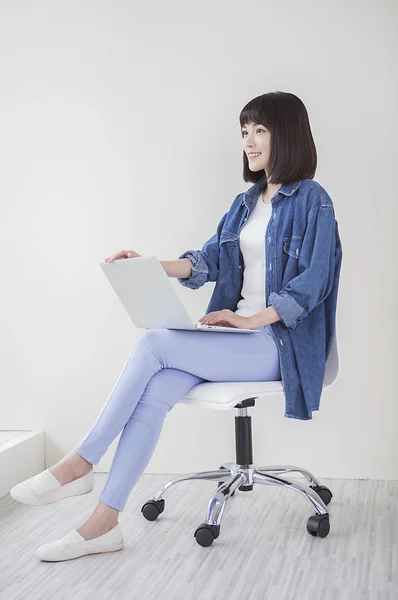 Young woman in a denim jacket sitting on a chair holding a laptop and smiling looking at it
