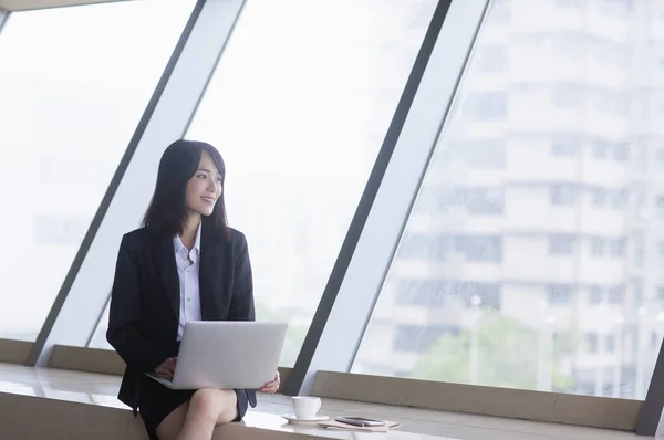 Young Asian woman in a suit using a laptop and smiling looking away,