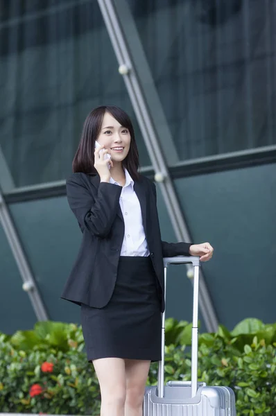 Young Asian woman in a suit holding a luggage and smiling talking on the phone