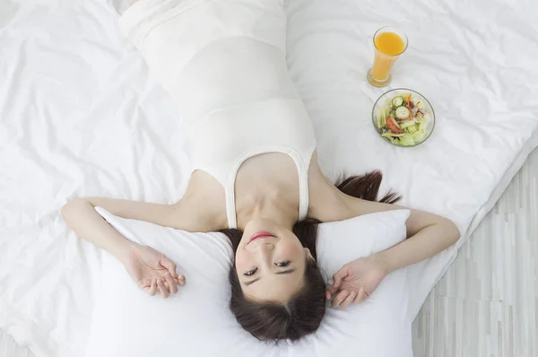 サラダ オレンジ ジュースの横にあるベッドの上に横たわるとカメラに笑顔の若いアジア女性 — ストック写真