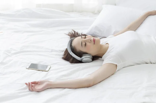 若いアジア女性ベッドに横になっていると 音楽に耳を傾け笑みを浮かべて — ストック写真
