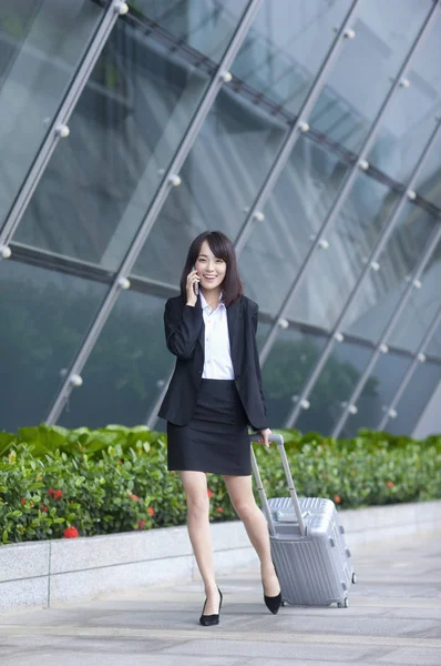 Young Asian woman in a suit holding a luggage and smiling talking on the phone
