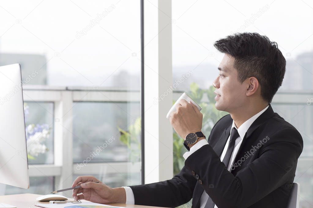 Young man in a suit sitting at the desk holding a cup of coffee and smiling looking away