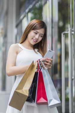 Bazı çanta tutarak ve cep telefonu kullanarak gülümseyen Genç Asyalı kadın