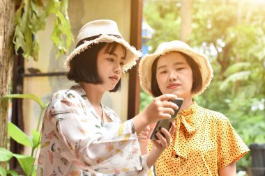 İki Asyalı kız arkadaş açık hava parkında internette gezerken cep telefonu kullanıyor. Jenerasyon dijital iletişim kavramı.