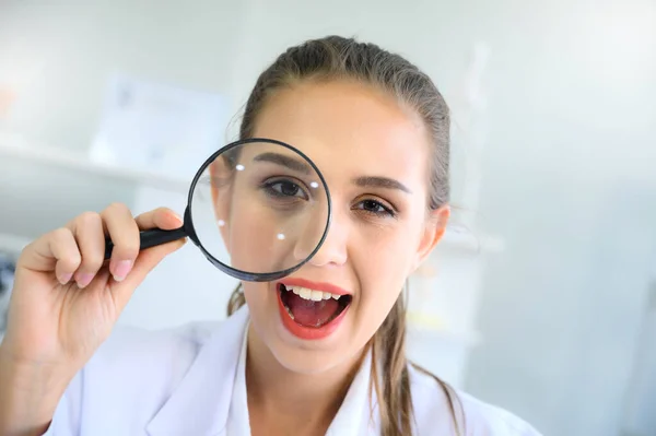 Cheerful beautiful young girl in white gown uniform looking at camera through magnifying glass. Close-up portrait shoot.
