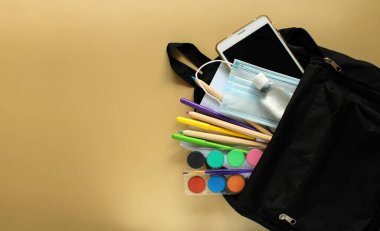 Kırtasiyeli okul çantasının üst görüntüsü: not defteri, kalemler, boyalar, işaretler, elektronik tablet, tıbbi maske ve sarı arka planda antiseptik. Okula geri dön ve koronavirüs ve diğer enfeksiyonlara karşı koruma sağla. Boşluğu kopyala