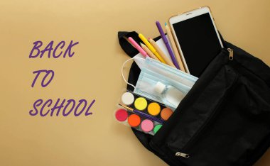 Kırtasiyeli okul çantasının üst görüntüsü: not defteri, kalemler, boyalar, işaretler, elektronik tablet, tıbbi maske ve bej arka planda antiseptik. Okula ve Coronavirus COVID-19 'a yattı. Menekşe keçeli kalemle okula geri dönüş imzası