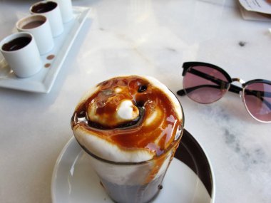 Krem şanti ve şerbetli taze yapılmış kahve içeceği mermer masanın üzerinde, şık güneş gözlüklerinin yanında berrak bir bardakta.        