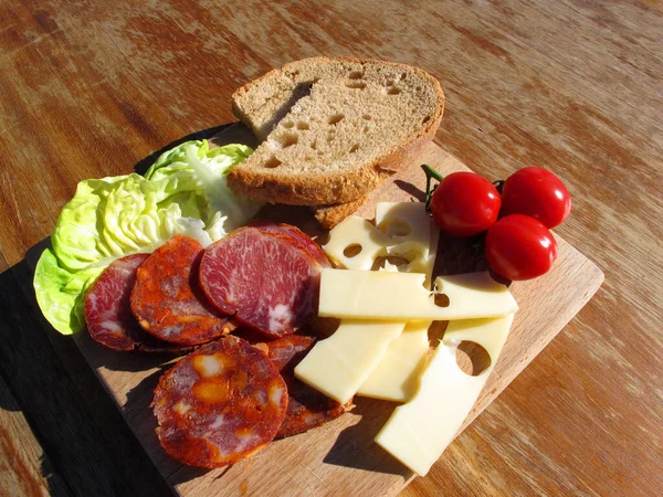 Ev yapımı sosis, salam, peynir, kiraz domatesleri, ahşap bir tahtanın üzerinde marul ve ekmek. Parlak güneş tarafından aydınlatılan eski ahşap bir masada basit ve lezzetli yemekler.                       