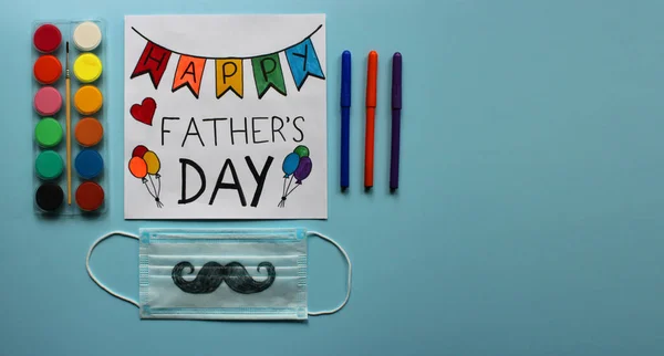 Kutlama kartı Mutlu Babalar Günü 'nde boyalar, keçeli kalemler ve baba için hediye olarak bir çocuğun çizdiği bıyıklı koruyucu yüz maskesi. Mavi arka plan. Koronavirüs salgını bağlamında Babalar Günü kavramı. Pankart