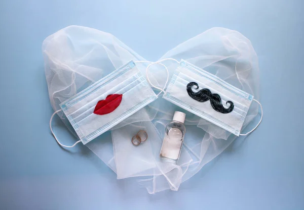 Two wedding rings, protective face masks with a mustache and lips painted on them and antiseptic are on blue background. The concept of wedding ceremonies during a pandemic of coronavirus COVID-19