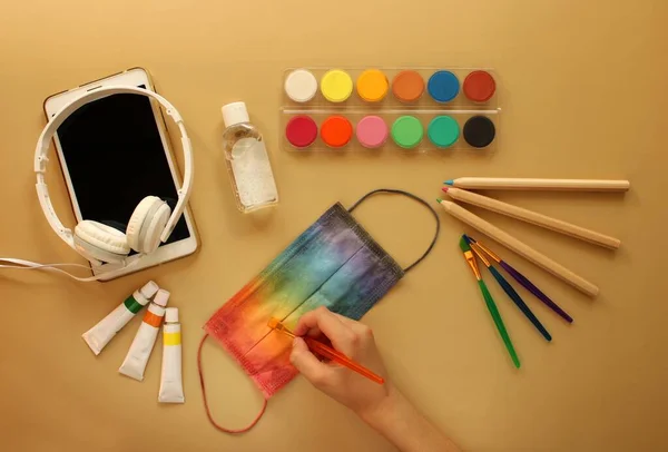 A child\'s hand paints a protective mask in rainbow colors next to school stationery, antiseptic, electronic tablet and headphones. Yellow background. Back to school and protection against coronavirus.