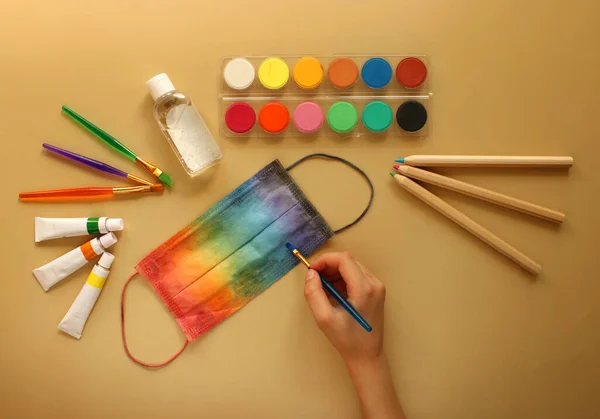 A child\'s hand paints a protective mask in rainbow colors next to school stationeries and antiseptic. Yellow background. Back to school and protection against coronavirus and other infections