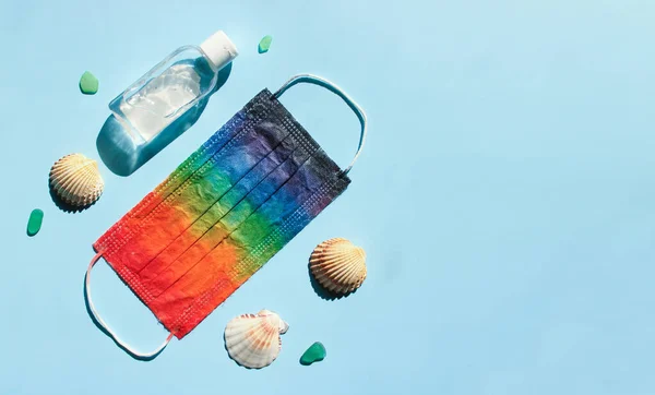 Yaz 2020 konsepti. Seyahat, tatil ve Coronavirus ve diğer enfeksiyonlara karşı koruma. Gökkuşağı renkleriyle boyanmış koruyucu maske, antiseptik ve güneş tarafından aydınlatılan mavi arka planda deniz kabukları. Boşluğu kopyala