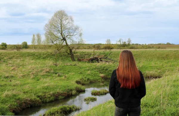 長い赤い耳を持つ少女は 明るい緑の草で覆われた小さな川のほとりに立って孤独な木の風景を賞賛します — ストック写真