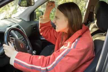 Kızıl saçlı kızgın genç kadın sürücü diğer sürücüleri yumruğuyla tehdit ediyor. Trafik sıkışıklığı konsepti. Stresli kadın sürücü arabasında oturuyor. 