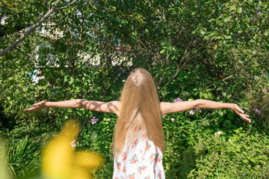 Altın saçlı kız kollarını açık yeşil yapraklara ve çimenlere doğru açtı. Mutlu çocukluk kavramı, güneşli yaz, sonbahar, hoş anılar ve duygular.