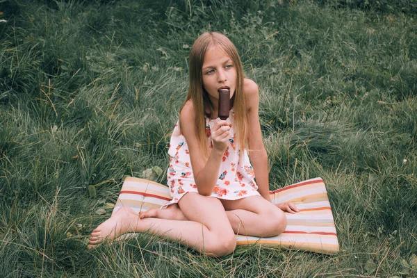 Altın saçlı kız açık yeşil çimenlerde oturur ve çikolatalı dondurma yer. Mutlu çocukluk, güneşli yaz, hoş anılar ve duygular. Dondurma Günü