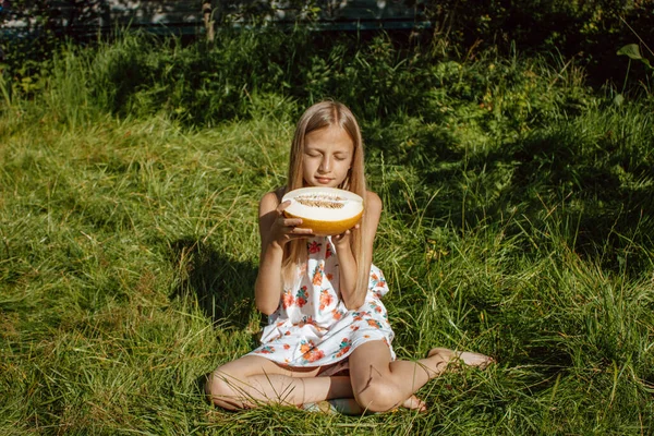 Altın saçlı kız bahçedeki yeşil ağaçların arka planında olgun kavun kokusunu içine çekiyor. Güneşli yaz, sonbahar, hasat, aroma, duygular, hoş anılar.