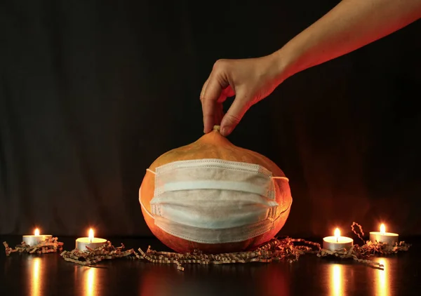 Annemin eli, Cadılar Bayramı balkabağını yanan mumların yanındaki masaya tek kullanımlık koruyucu bir maskenin içine koyar. Siyah arka plan. Coronavirus salgını sırasında Cadılar Bayramı partisi için ev dekorasyonu.