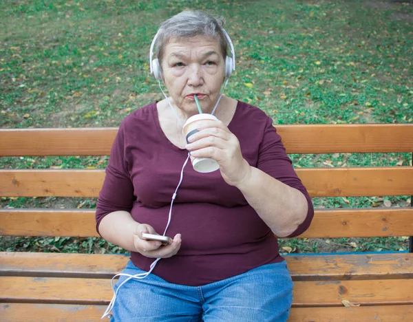 Elderly woman sits on bench in the yard with phone, headphones and cup of coffee and listens to music. The concept of  adaptation of older people in modern life. International Day of Older Persons and Grandparents Day