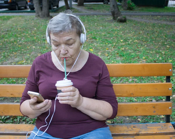 Yaşlı bir kadın telefon, kulaklık ve kahveyle bahçede bankta oturur ve müzik dinler. Modern hayatta yaşlı insanların uyum sağlama kavramı. Uluslararası Yaşlılar ve Büyükbabalar Günü