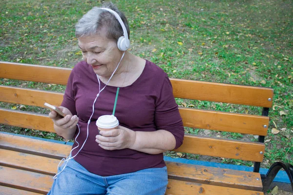 Elderly woman with headphones and cup of coffee sits on bench and talks on the phone with her family. The concept of  adaptation of older people in modern life. International Day of Older Persons and Grandparents Day