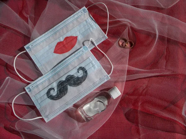 Bıyıklı ve dudakları boyanmış iki tek kullanımlık yüz maskesi, kırmızı arka planda alyans ve antiseptik var. Coronavirus COVID-19 salgınında düğün seremonisi konsepti
