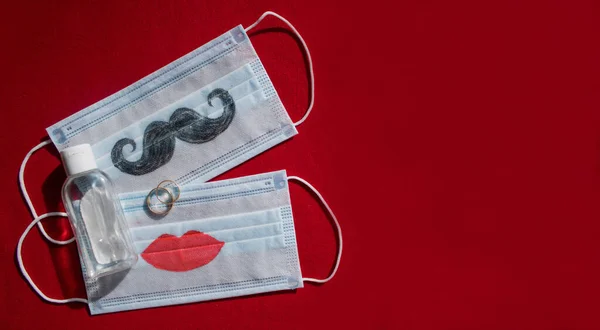 Bıyıklı ve dudakları boyanmış iki tek kullanımlık yüz maskesi, kırmızı arka planda alyans ve antiseptik var. Coronavirus COVID-19 salgınında düğün seremonisi konsepti