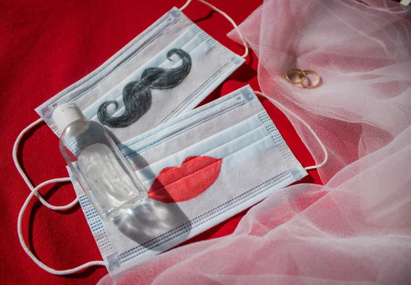 Bıyıklı ve dudakları boyanmış iki tek kullanımlık yüz maskesi, kırmızı arka planda alyans ve antiseptik var. Coronavirus COVID-19 salgınında düğün seremonisi konsepti