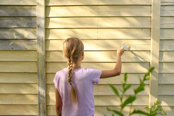 Saç örgülü, sarı saçlı reşit olmayan bir kız bir yazlık evin duvarını fırçayla sarıya boyuyor. Ev tadilatı ve aile kavramına yardım. Yaşam biçimi