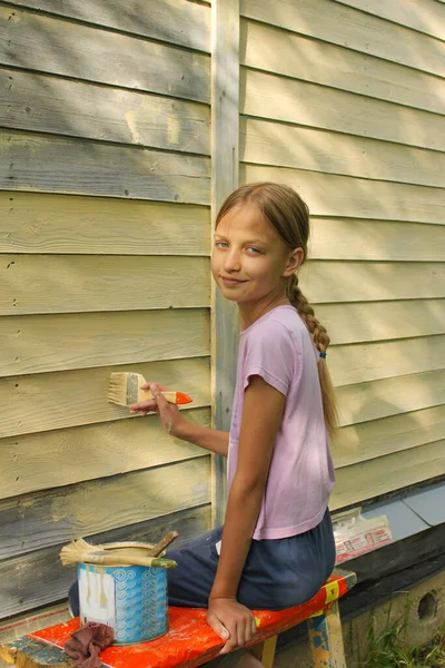 Saç örgülü, sarı saçlı reşit olmayan bir kız bir yazlık evin duvarını fırçayla sarıya boyuyor. Ev tadilatı ve aile kavramına yardım. Yaşam biçimi
